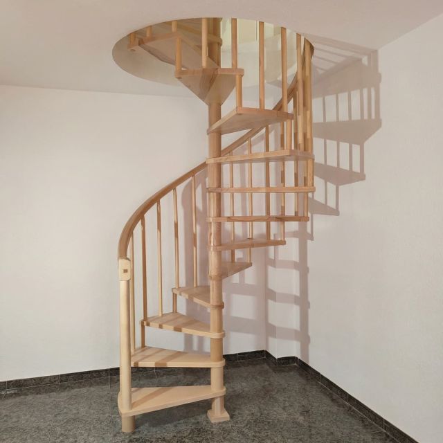 Una elegante escalera de madera caracol de tono claro y diseño helicoidal, instalada en el rincón de una estancia minimalista con paredes blancas y suelo de baldosas oscuras.
