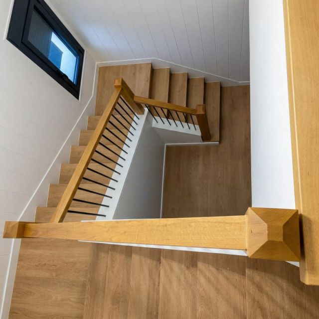 Vista cenital de una escalera en u con peldaños de madera clara y barandilla de finos listones negros, que conecta los niveles de una vivienda de estilo contemporáneo y paredes blancas.