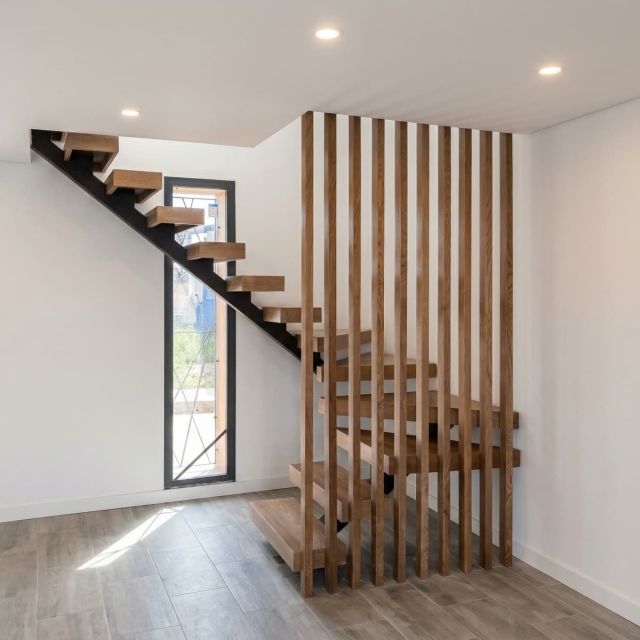 Diseño de un cerramiento de escalera moderno compuesto por una celosía de listones de madera verticales que nacen desde el suelo, junto a una escalera volada con peldaños de madera y estructura metálica negra en un ambiente de paredes blancas.