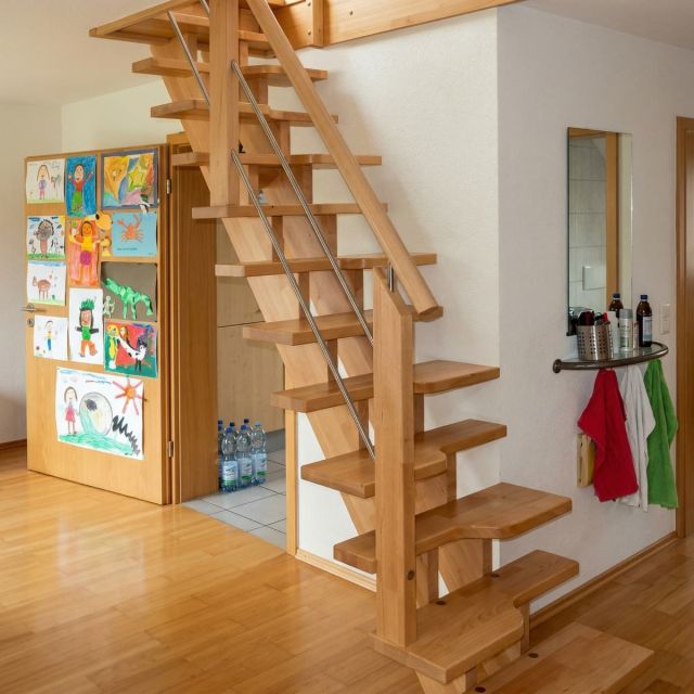 Una escalera de pasos alternados fabricada en madera clara con pasamanos metálico, diseñada para maximizar el ahorro de espacio en un interior doméstico con acabados en madera.